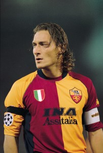 Totti, ídolo da Roma, com a tradicional camisa amarela e vermelha. Escudo traz a loba capitolina.